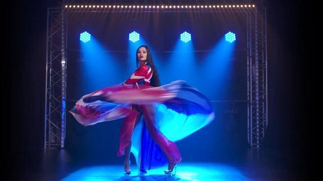 Slender high girl dancing in a sexy suit in the color of the English flag and rhinestones. Attractive brunette waving her skirt in a dark studio with smoke and blue neon lights.