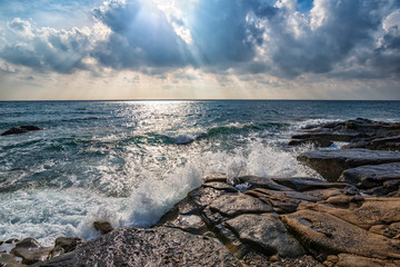 Sea waves break on a wild rocky shore under a dramatic sky with sunbeams through the clouds