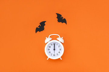 White alarm clock and black cardboard bats on orange background. Halloween minimal concept. Top...