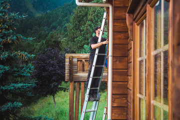 Man on a ladder working on a wooden cottage with trees and mountains behind