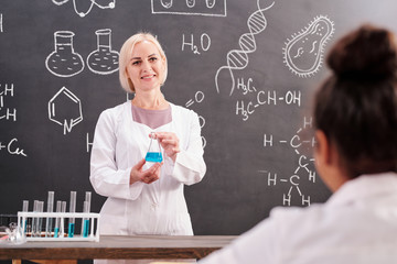 Young blond smiling teacher of chemistry holding tube with blue liquid substance