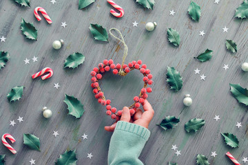 Heart wreath decorated with frosted berries. Geometric Christmas background decorated with holly leaves, red white stripy candy canes and paper stars. Top view on aged wood.