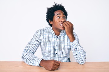 Handsome african american man with afro hair wearing casual clothes sitting on the table looking stressed and nervous with hands on mouth biting nails. anxiety problem.