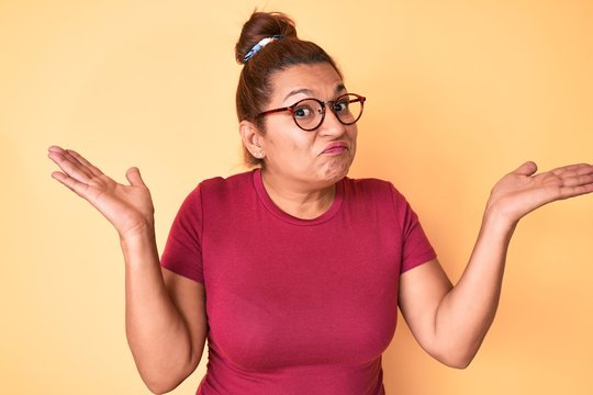 Middle age brunette hispanic woman wearing casual clothes and glasses clueless and confused with open arms, no idea and doubtful face.