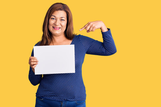 Middle age latin woman holding paper banner with blank space pointing finger to one self smiling happy and proud