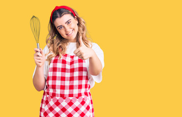 Young caucasian woman wearing apron holding whisk smiling happy and positive, thumb up doing excellent and approval sign