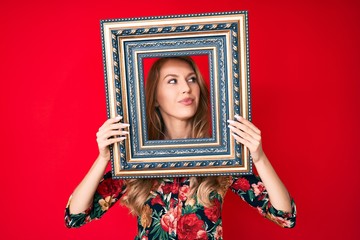 Young caucasian woman with blond hair holding empty frame smiling looking to the side and staring away thinking.