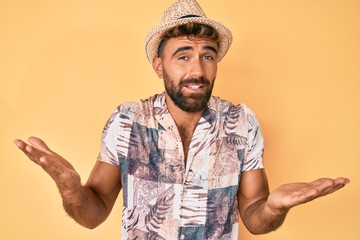 Young hispanic man wearing summer hat clueless and confused expression with arms and hands raised....
