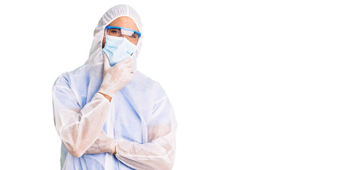 Young hispanic man wearing doctor protection coronavirus uniform and medical mask looking confident at the camera smiling with crossed arms and hand raised on chin. thinking positive.