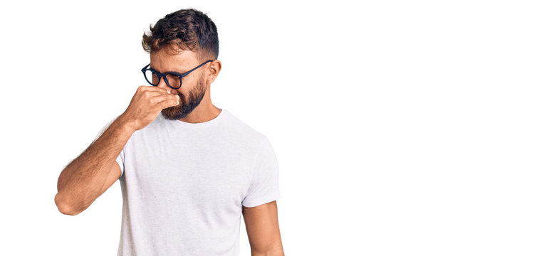 Young hispanic man wearing casual clothes and glasses smelling something stinky and disgusting, intolerable smell, holding breath with fingers on nose. bad smell
