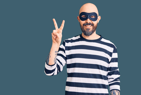 Young handsome man wearing burglar mask smiling looking to the camera showing fingers doing victory sign. number two.