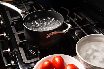 Black pot with water boiling on black stove with white bowl and red tomatoes