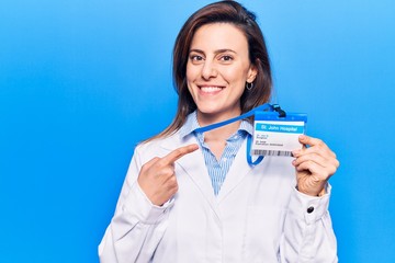Young beautiful woman wearing doctor stethoscope holding id card smiling happy pointing with hand...