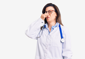 Young beautiful woman wearing doctor stethoscope and glasses smelling something stinky and disgusting, intolerable smell, holding breath with fingers on nose. bad smell