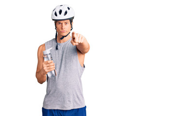 Young handsome man wearing bike helmet holding bottle of water pointing with finger to the camera and to you, confident gesture looking serious