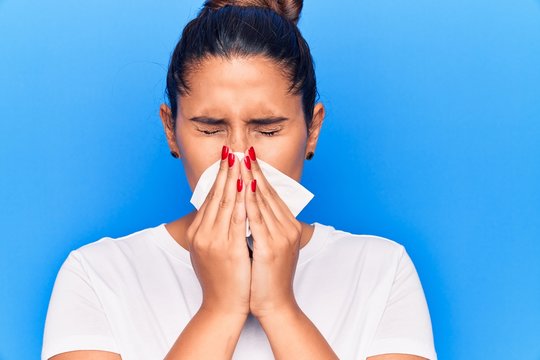 Young latin woman illness using paper handkerchief on nose. Standing over isoltated blue background