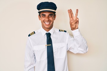 Young hispanic man wearing airplane pilot uniform showing and pointing up with fingers number three while smiling confident and happy.