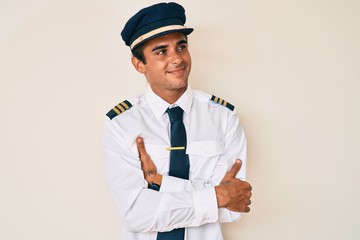 Young hispanic man wearing airplane pilot uniform looking to the side with arms crossed convinced and confident