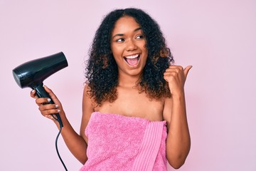 Young african american woman wearing shower towel holding dryer pointing thumb up to the side smiling happy with open mouth