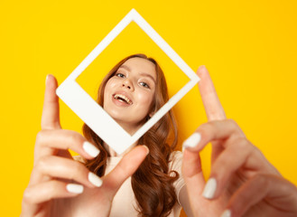 young smiling woman holding in hands photo frame with her face isolated on yellow background,...