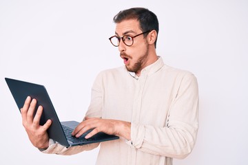Young handsome man holding laptop scared and amazed with open mouth for surprise, disbelief face