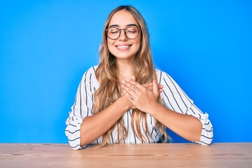 Young beautiful blonde woman wearing glasses sitting on the table smiling with hands on chest with closed eyes and grateful gesture on face. health concept.