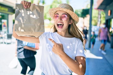 Young beautiful blonde caucasian woman smiling happy outdoors on a sunny day pointing to delivery paper bag
