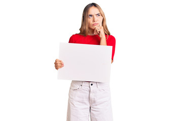 Naklejka premium Young beautiful blonde woman holding blank empty banner serious face thinking about question with hand on chin, thoughtful about confusing idea