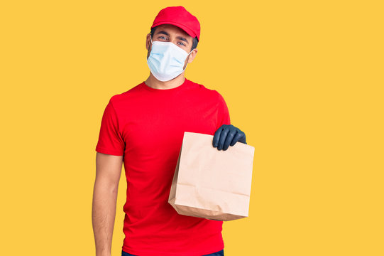 Young hispanic man delivering food wearing covid-19 safety mask holding paper bag looking positive and happy standing and smiling with a confident smile showing teeth