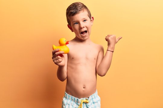 Cute blond kid wearing swimwear holding duck toy pointing thumb up to the side smiling happy with open mouth