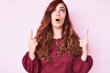 Young beautiful woman wearing casual winter sweater amazed and surprised looking up and pointing with fingers and raised arms.