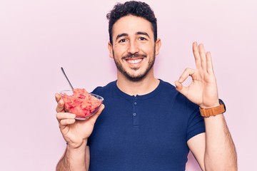 Young hispanic man holding ice cream doing ok sign with fingers, smiling friendly gesturing...