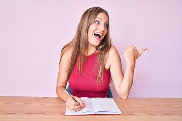 Young caucasian girl writing notebook sitting on the table pointing thumb up to the side smiling happy with open mouth