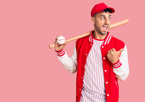 Young hispanic man playing baseball holding bat and ball pointing thumb up to the side smiling happy with open mouth