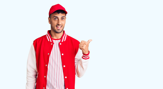 Young hispanic man wearing baseball uniform smiling with happy face looking and pointing to the side with thumb up.