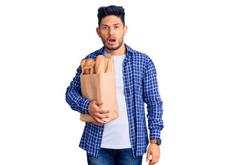 Handsome latin american young man holding paper bag with bread in shock face, looking skeptical and...