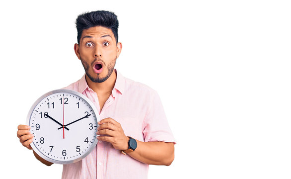 Handsome latin american young man holding big clock scared and amazed with open mouth for surprise, disbelief face