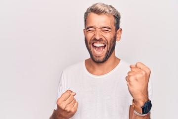 Young handsome blond man wearing casual t-shirt standing over isolated white background celebrating surprised and amazed for success with arms raised and eyes closed