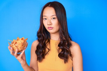 Young beautiful chinese girl holding nachos potato chips thinking attitude and sober expression looking self confident