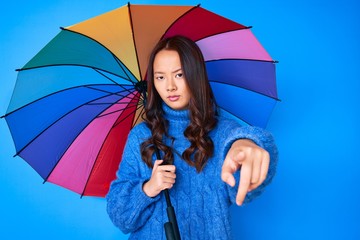 Young beautiful chinese girl holding colorful umbrella pointing with finger to the camera and to you, confident gesture looking serious