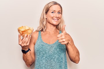 Middle age caucasian blonde woman holding potato chips smiling happy and positive, thumb up doing...