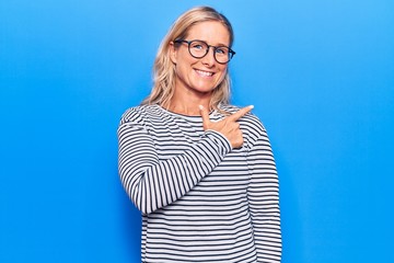 Obraz na płótnie Canvas Middle age caucasian blonde woman wearing casual striped sweater and glasses cheerful with a smile of face pointing with hand and finger up to the side with happy and natural expression on face