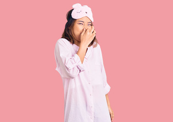 Young beautiful chinese girl wearing sleep mask and pajama smelling something stinky and...
