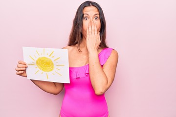 Young beautiful brunette woman wearing pink swimwear holding paper with sun draw covering mouth with hand, shocked and afraid for mistake. Surprised expression