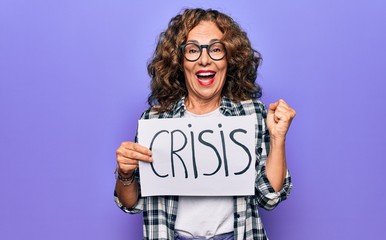 Middle age beautiful woman holding paper with crisis message over purple background screaming proud, celebrating victory and success very excited with raised arm