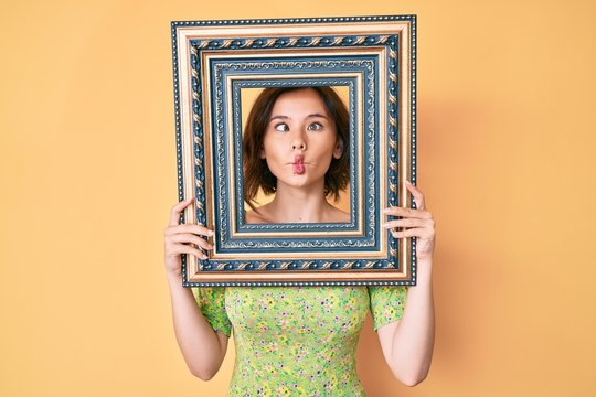 Young beautiful woman holding empty frame making fish face with mouth and squinting eyes, crazy and comical.