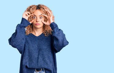Young blonde woman with curly hair wearing casual winter sweater trying to open eyes with fingers,...