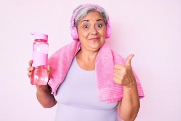 Senior hispanic woman wearing gym clothes and using headphones holding bottle of water smiling happy and positive, thumb up doing excellent and approval sign