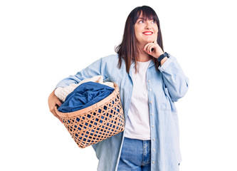 Young plus size woman holding laundry basket serious face thinking about question with hand on...