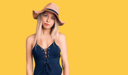 Young beautiful blonde woman wearing swimwear and summer hat with serious expression on face. simple and natural looking at the camera.